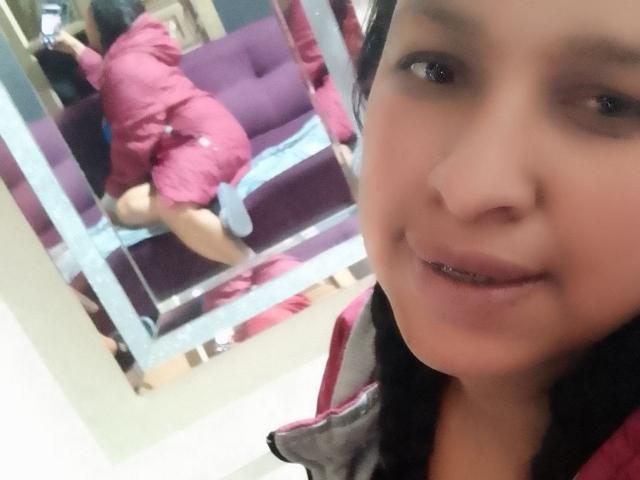 ZoehFerraro in pink dress, kneeling on purple couch, reflected in mirror. Foreground: ZoehFerraro's smiling face, black hair, and visible teeth. Mirror shows her ass and back.