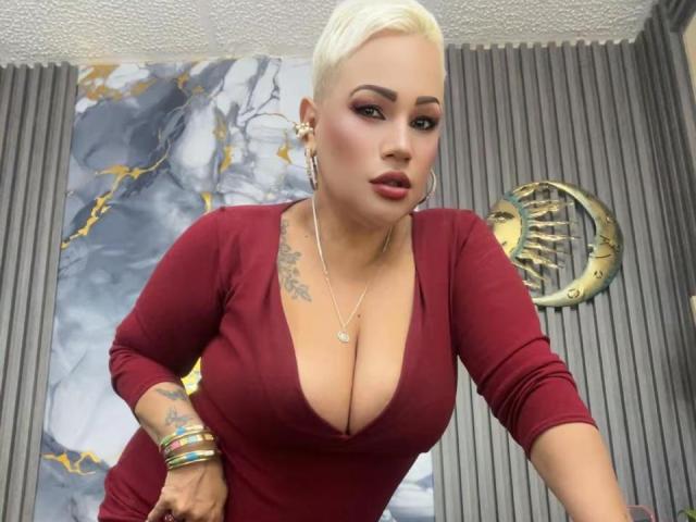 GianellaCox with platinum blonde hair, deep red dress, showing large tits, tattoo on left arm, gold hoop earrings, multiple bracelets, and a serious expression.
