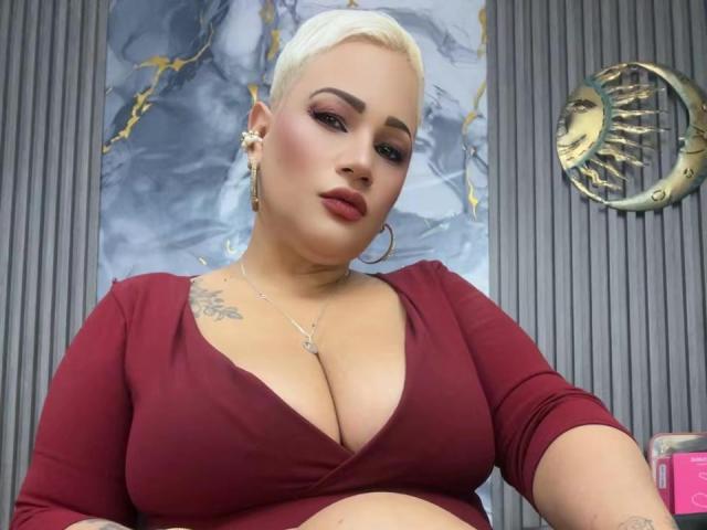 GianellaCox, avec une coupe de cheveux pixie blonde, de grandes seins, une robe rouge vif, des boucles d'oreilles pendantes, un tatouage sur la poitrine gauche, du rouge à lèvres rouge et un maquillage discret, debout devant un mur gris à nervures..