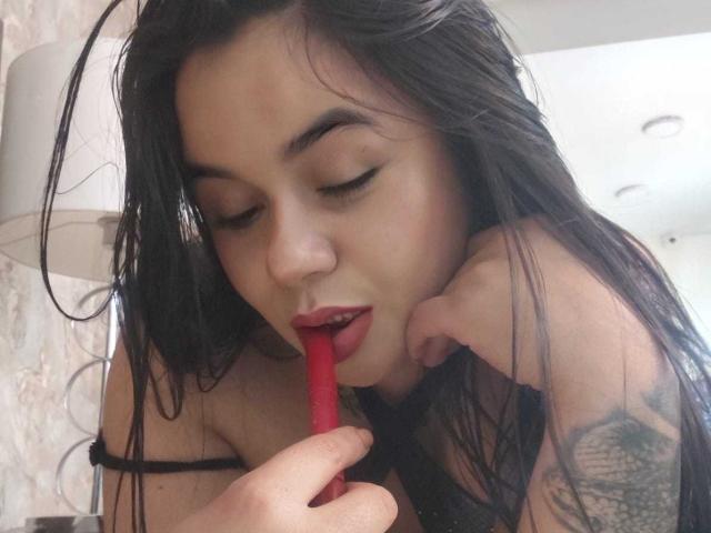 EveWillis with wet black hair, red lipstick, and tattoo on arm, sucking on a red lipstick tube, eyes closed. She has medium tits and a cute face.