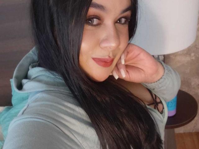 EveWillis with long black hair, wearing a green hoodie, resting her chin on her hand, showing her red lipstick and small tits, smiling at the camera.