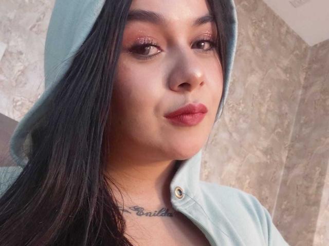 EveWillis with long black hair, wearing a light blue hoodie, has glossy pink lipstick, shimmering brown eyes, and a tattoo under her chin. Her face is centered in the shot.