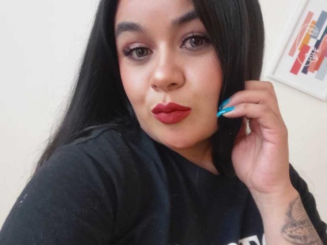 EveWillis has long black hair, red lipstick, and blue nail polish, with a tattoo on her right arm, wearing a black shirt, and giving a pouty look.