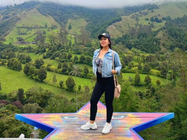 Katty Rivera è in piedi su una piattaforma di legno a forma di stella, indossa una giacca blu, leggings neri, scarpe da ginnastica bianche e un cappello, tiene una borsa beige, con montagne verdi sullo sfondo..