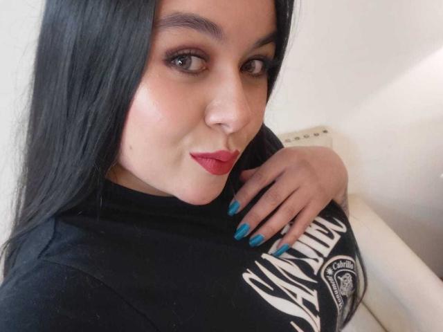 EveWillis with long black hair, red lipstick, and blue nails, holding her black shirt over her tits, looking at the camera with a slight smile.