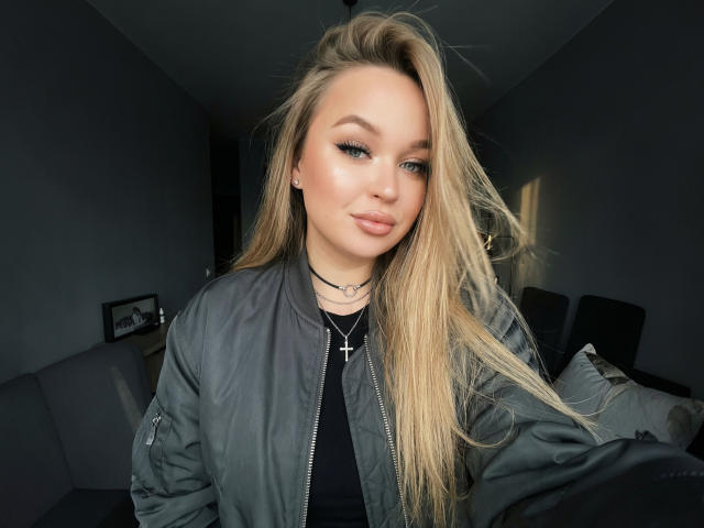 Velorda with long blonde hair, wearing a grey bomber jacket and black top, has blue eyes and full pink lips. Cross necklace visible. She's looking at the camera.