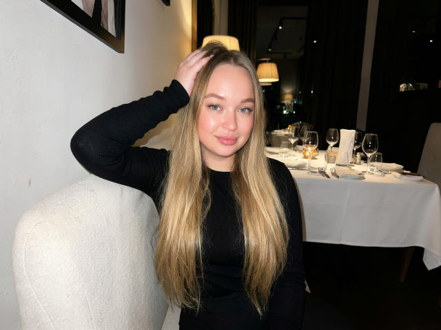 Velorda with long blonde hair, wearing a black long-sleeve top, sitting on a white chair, hand in hair, smiling at camera, table with glasses and candles in background.