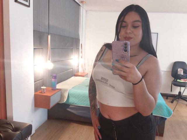 EveWillis with long black hair, wearing a white crop top, showing her tits, and black pants, taking a mirror selfie in a bedroom. Her phone has a pink case.