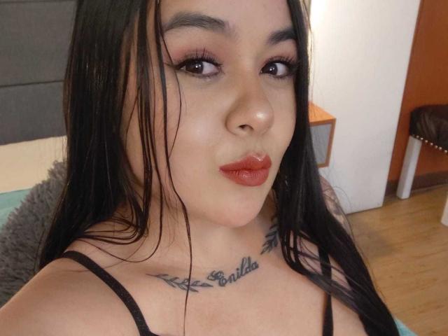 EveWillis with long black hair, wearing red lipstick, eyeliner, and black spaghetti straps, showing a tattoo under her chin that reads "Beautiful". Her ass is partially visible.
