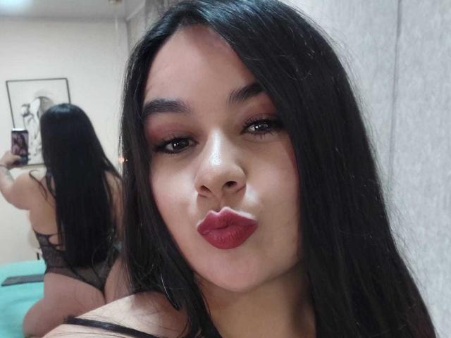 EveWillis with long black hair, red lipstick, and brown eyes, pouting her lips, wearing a black lace bra, reflected in a mirror showing her back and butt.