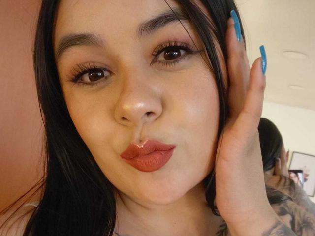 EveWillis has black hair, blue nails, and a thick white stream on her pouty red lips, with her hand touching her cheek. She has tattoos on her visible arm.