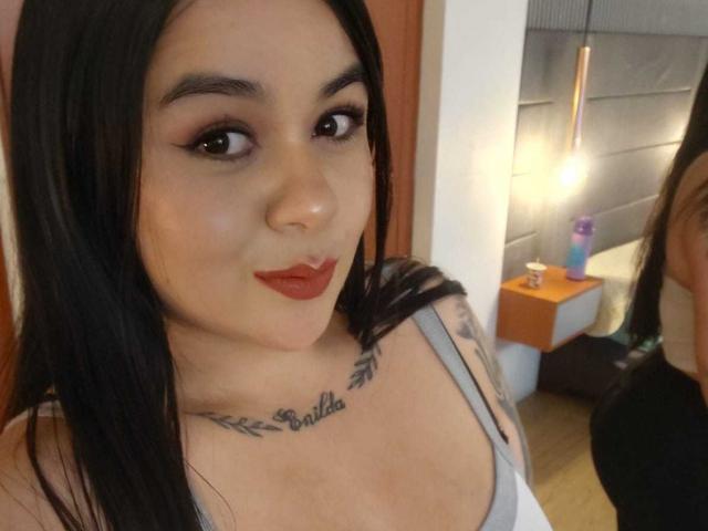 EveWillis with black hair, brown eyes, red lipstick, gray tank top, tattoo under chin, mirror reflection showing her back, small round nose, light skin, in a bedroom.