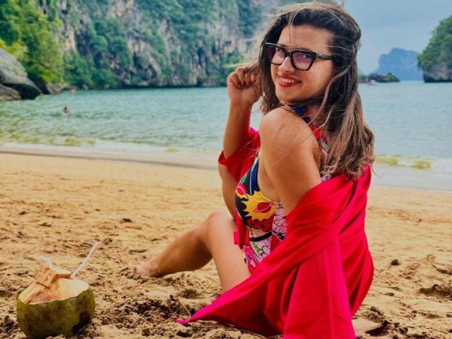 BustyMira with glasses, red shawl, colorful floral swimsuit, sitting on sandy beach, smiling, brown hair, coconut with straw, green cliffs in background, water, sunny day.
