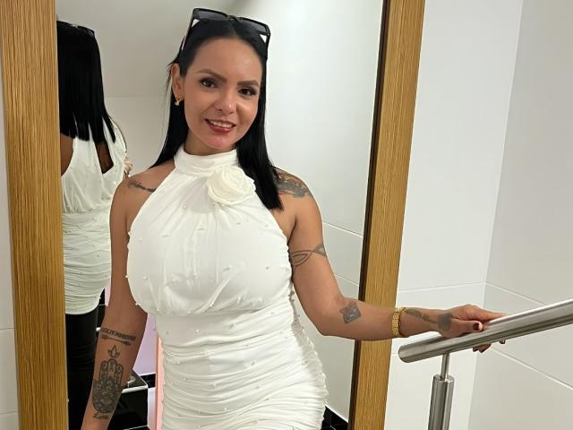 JessicaFiore, avec ses cheveux noirs, portant une robe blanche à encolure dégagée, debout près d'un miroir, tenant une rambarde, montrant des tatouages sur ses bras, souriante, avec des lunettes de soleil sur la tête..