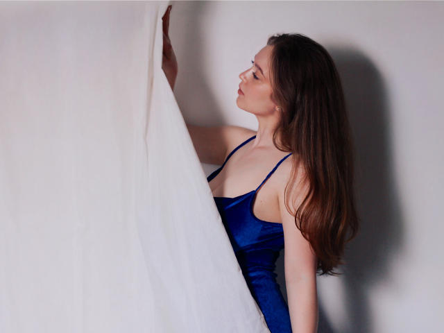 ValeriVoom, avec ses longs cheveux bruns, portant une robe en satin bleu, tient des rideaux blancs, et regarde sur le côté avec une expression pensive. Ses seins sont petits, et ses fesses ne sont pas visibles..