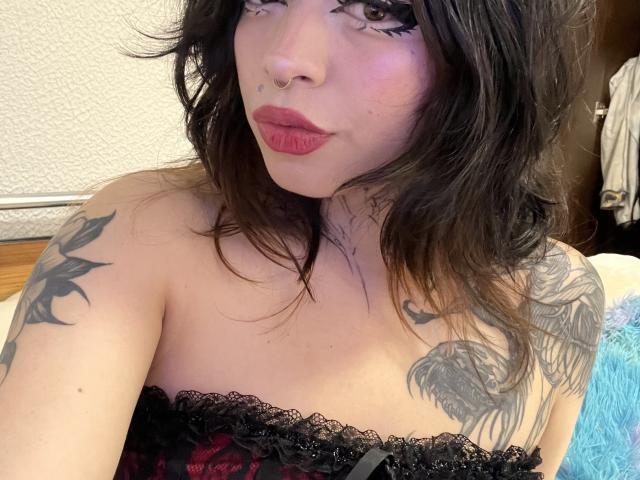 BabeSix with wavy brown hair, red lipstick, black lace strap dress, tattoos on shoulders, nose ring, and thick black eyeliner, looking at camera with a slight pout.