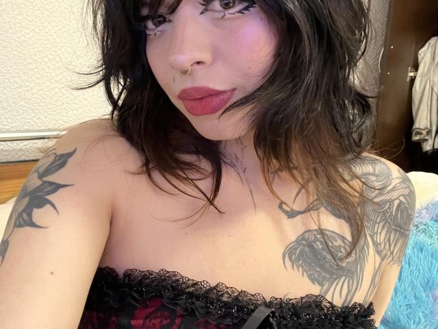 BabeSix with dark, wavy hair, nose ring, and red lipstick, showing tattoos on shoulders, wearing black lace strapless top, looking at camera with slightly pouty expression.