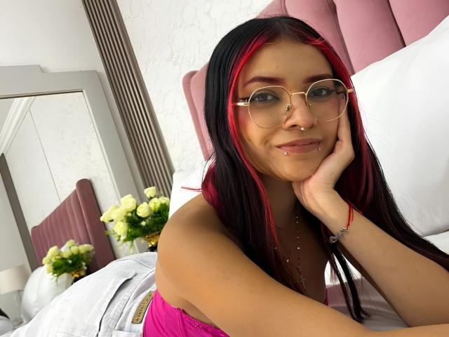 AriaRosel with red-black hair, glasses, nose ring, and lip piercing, resting on pink bed, wearing pink top, smiling, hand on cheek, white walls, flowers in background.