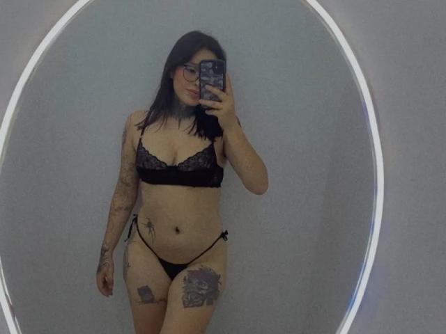 PaisleyMour con reggiseno e perizoma neri, che mostra tatuaggi sulle braccia e sulla coscia, tiene in mano un telefono, di fronte a uno specchio circolare con una cornice bianca. Il suo seno è piccolo, i glutei non sono visibili..