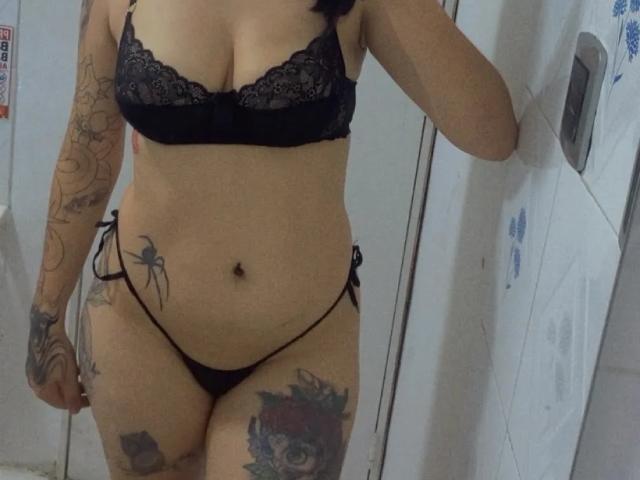 PaisleyMour usando um sutiã e tanga de renda preta, mostrando tatuagens nos braços e coxas, com o umbigo visível e uma pequena tatuagem de aranha no lado direito. A renda transparente cobre seus seios..