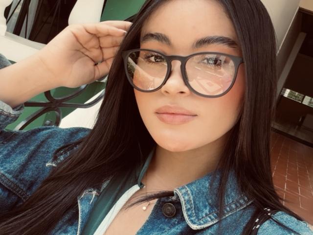 MiiaPalmer with long black hair, wearing black-framed glasses, a blue denim jacket, and a green strap, looking at the camera with a slight smile.
