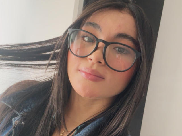 MiiaPalmer with dark, straight hair, wearing large black glasses, a blue denim shirt, and pinkish lipstick, has a small red mark on her right cheek.