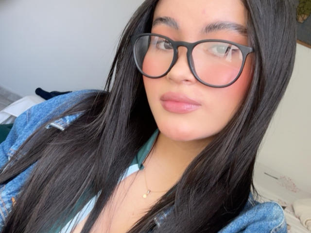 MiiaPalmer with long black hair, wearing black glasses and a blue denim jacket, showing her pink lips and light skin, has a small gold necklace and a cute face.