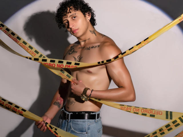 Shirtless JayTaylorr with curly black hair, tattoos, and a bracelet, holding yellow crime tape across his chest and belly. Wearing blue jeans, standing against a shadowy wall.