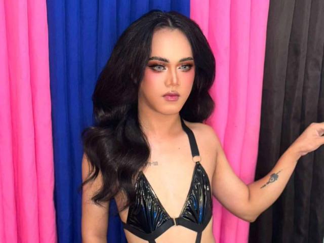 GorgeousShanelxx, avec ses longs cheveux noirs, porte un soutien-gorge en vinyle noir brillant, a un tatouage sur son bras droit, et il y a un rideau rose et bleu derrière elle..