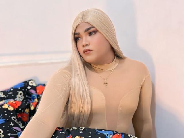 Aica, deliciosa, con cabello rubio largo, busto generoso, blusa beige transparente, collar con cruz, expresión de labios fruncidos, sentada en una cama colorida, mirando hacia un lado, con iluminación suave..