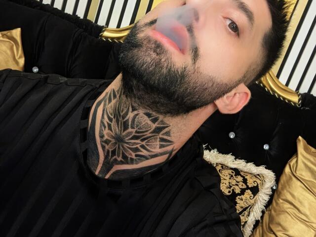 TommyDum with a black striped shirt, beard, and intricate tattoo on his neck. Blurred face, short dark hair, lying on black and gold pillows.