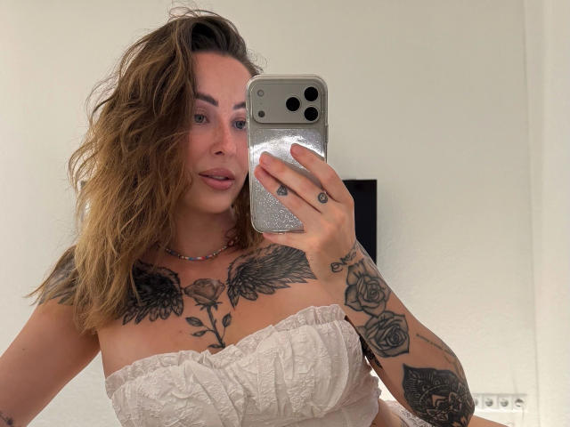 Meltattt-hot with wavy brown hair, white strapless top, tattoos on chest and arms, holding phone, pink lips, wearing necklace, taking mirror selfie, showing tits and rose tattoos.