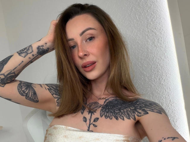 Meltattt-hot with long brown hair, tattoos on arms and chest, wearing a strapless white top, looking at camera with hand in hair, pink lips, light freckles.