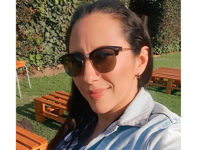 Jade Foster, avec ses lunettes de soleil foncées, portant une chemise en jean et affichant un léger sourire, a de longs cheveux tressés et de petites boucles d'oreilles, et se tient dans un cadre extérieur ensoleillé avec des bancs en bois..