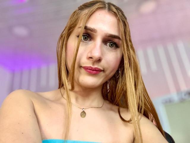 AndyDupont with wet blonde hair, green eyes, pink lipstick, bare shoulders, blue top, small necklace, smiling slightly, fair skin, soft light, slight purple glow in background.