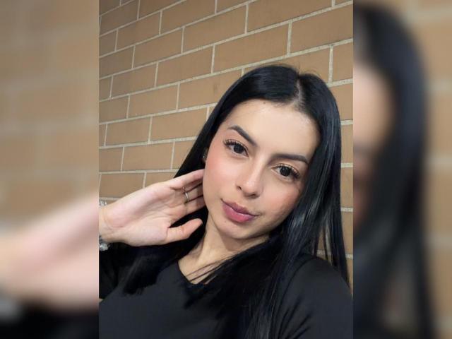 Isabella James tiene el pelo largo y negro, lápiz labial rosa, y está tocándose la mejilla con una mano. Lleva una blusa negra y tiene una ligera sonrisa. Espejo reflectante a la derecha..