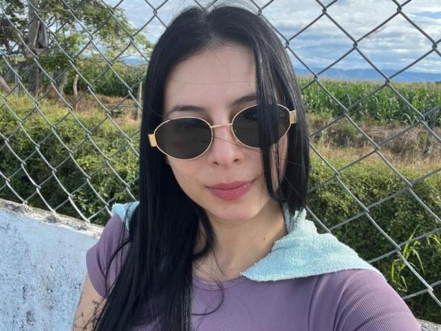 IsabellaJames has long black hair, wearing round sunglasses with gold frames, a purple shirt with a light blue collar, and is standing in front of a chain-link fence.