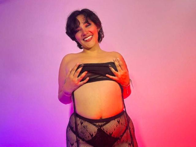 ChloeSterling with short dark hair, smiling, holding black strap above her tits, wearing black mesh panties, exposing her belly, standing against pink and purple gradient background.
