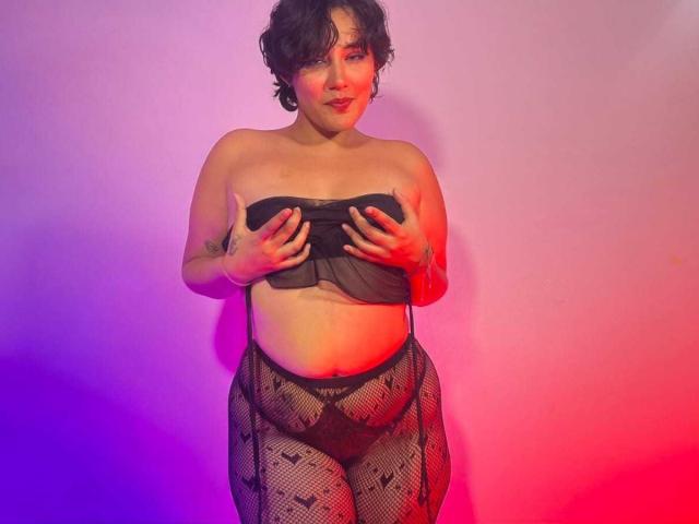 ChloeSterling with curly dark hair, fair skin, small tits, covering black mesh bra with hands, wearing fishnet pants, red and purple light on her body, slight smile.