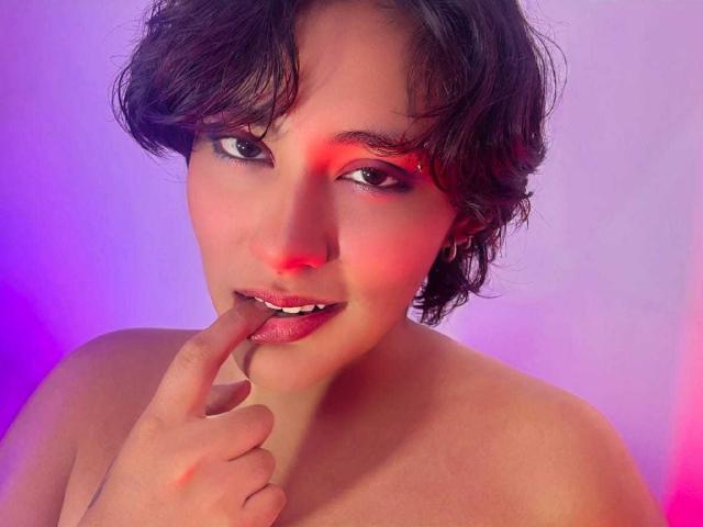 Chloe Sterling, avec ses courts cheveux noirs en bataille, touche ses lèvres rosées avec son doigt, nue, regardant de manière séduisante l'appareil photo, avec une lumière rouge et violette sur son visage..