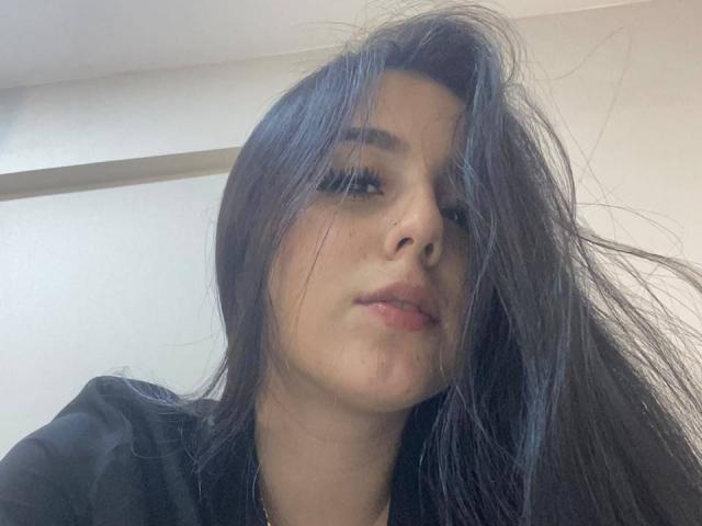 StefanyDelly, con i capelli neri spettinati che le coprono il viso, un trucco leggero, una camicia scura e un sorriso appena accennato, quasi impercettibile. I suoi occhi sono socchiusi e guardano leggermente verso il basso..