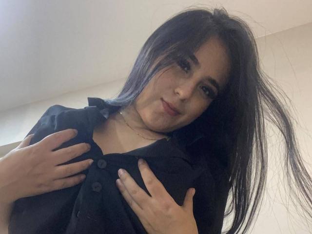 StefanyDelly with long black hair, wearing a black shirt, hands on her tits, slight smile, light skin, subtle makeup, necklace, taking a selfie from a low angle.