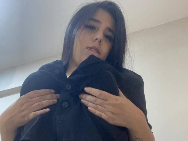 StefanyDelly with long dark hair, wearing a black shirt, holding her large tits with both hands, looking down at the camera with a slightly open mouth.