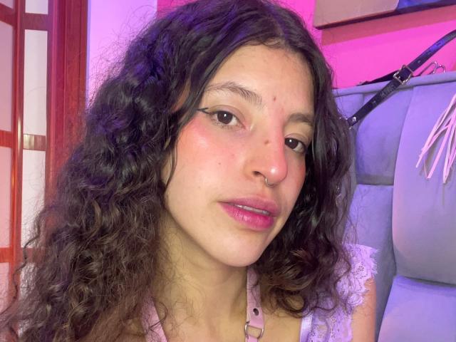 NamySex heeft krullend bruin haar, roze lippen, een neusring en witte eyeliner. Ze draagt een lichtroze top, met een achtergrond in paars en roze..
