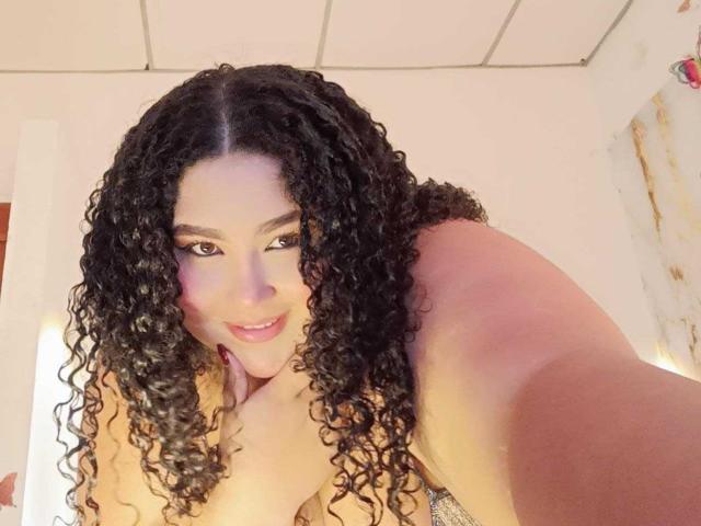 HurrenMatt with long curly black hair, smiling, topless, showing small tits, pink lipstick, tan skin, looking up at the camera, in a well-lit room.