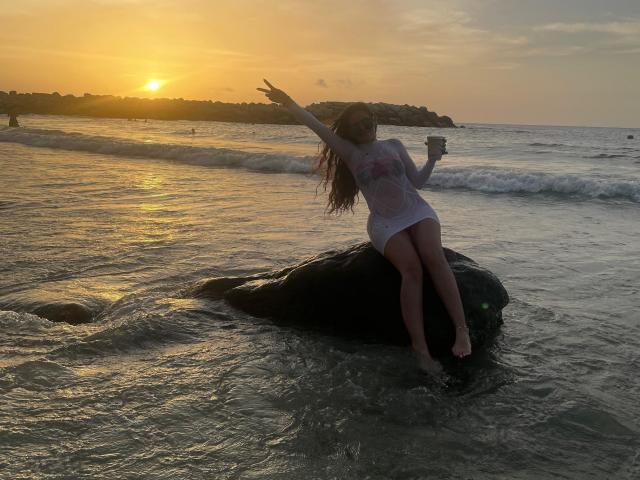 MoreForMyPussy with long brown hair, white wet dress, pointing up, sitting on black rock in shallow water at sunset, holding phone, small tits, sexy, ocean, rocks in background.