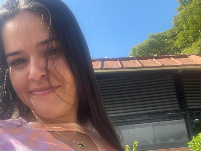 MoreForMyPussy has wet dark brown hair, a cute face, and a subtle smile. She's wearing a pink shirt and a necklace with a small pendant. Sunny blue sky.
