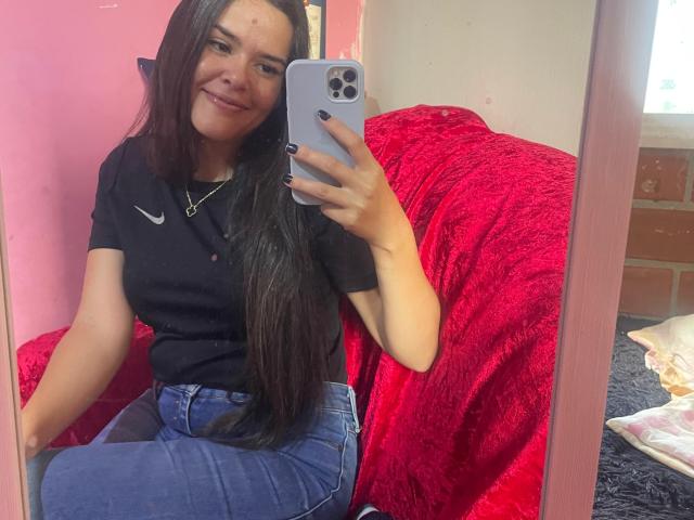 MoreForMyPussy with long black hair, smiling, wearing a black Nike polo and blue jeans, taking a mirror selfie with a white iPhone, red textured blanket behind her.