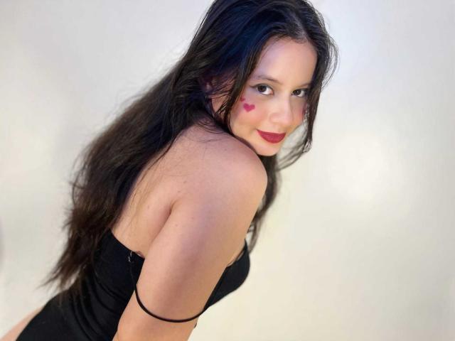 LunaRomano, con i suoi lunghi capelli neri, indossa un abito nero senza spalline, ha tatuaggi a forma di cuore rossi sulle guance, guarda indietro con un sorriso e rossetto rosso, e una fascia nera al braccio..