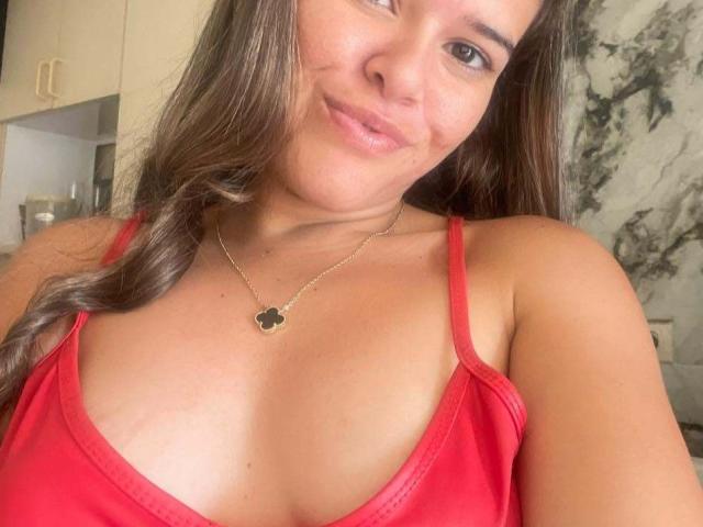 MoreForMyPussy with long brown hair, smiling, wearing a red tank top, gold necklace with a black flower, showing her boobs. Her skin is tan, she's cute.